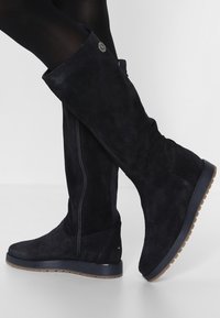 Botas altas de ante negro con suela plana, que cuentan con un cierre de cremallera lateral y un sutil detalle del logo cerca de la parte superior. Textura suave, forma ajustada.