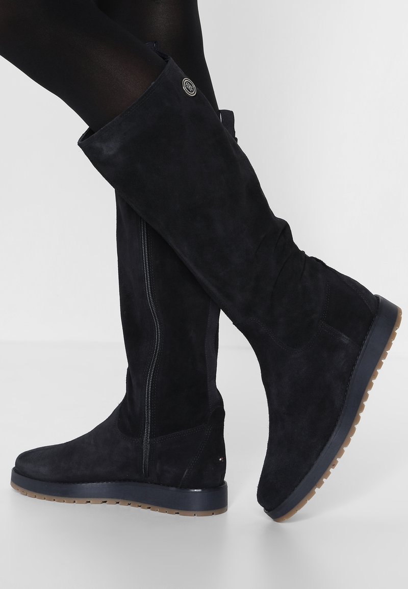 Botas altas de ante negro con suela plana, que cuentan con un cierre de cremallera lateral y un sutil detalle del logo cerca de la parte superior. Textura suave, forma ajustada.