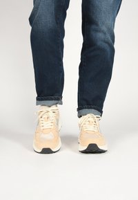 Lightweight sneakers in cream and peach, featuring mesh panels, laces, and a contrasting black outsole. Worn with cuffed dark jeans.