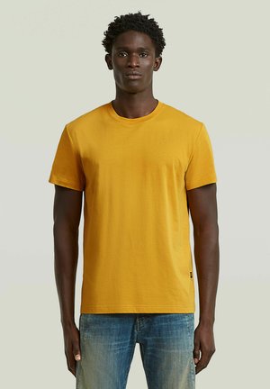 Hombre con camiseta lisa de manga corta color mostaza y vaqueros azules, de pie frente a un fondo claro y neutro.
