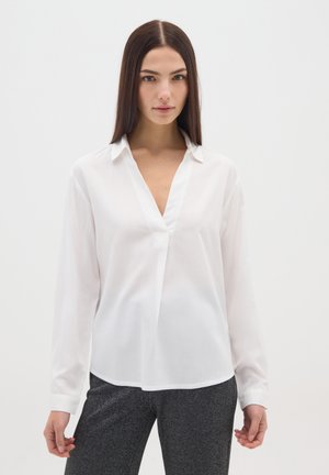 Jeune femme aux cheveux longs et foncés, portant un chemisier blanc à col en V et un pantalon gris foncé, se tenant devant un fond clair uni.