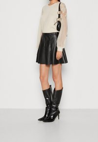 Cremefarbener Pullover mit transparenten, plissierten Ärmeln, kombiniert mit einem schwarzen, ausgestellten Lederrock und schwarzen kniehohen Stiefeln mit spitzer Form. Schwarze Handtasche inklusive.