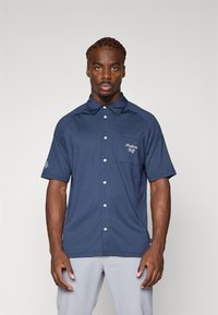 Kortermet marineblå polo skjorte med teksturert stoff, knappestolpe og brystlomme som har "Palabon Golf" brodert i hvitt.