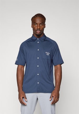 Kortermet marineblå polo skjorte med teksturert stoff, knappestolpe og brystlomme som har "Palabon Golf" brodert i hvitt.