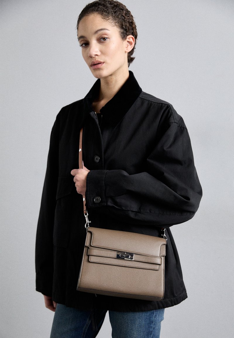 Sac à main en cuir texturé marron avec un rabat à l'avant et des accessoires en métal argenté, porté en bandoulière, associé à une veste noire et un jean bleu.