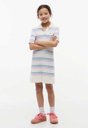 Ragazza sorridente con le braccia incrociate, che indossa un vestito di maglia bianco con strisce pastello, calzini bianchi e scarpe da ginnastica rosa su sfondo bianco.