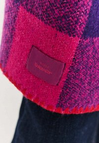 Dzianinowy szalik w żywym różowo-fioletowym wzorze w kratę. Miękka faktura z przeszytym brzegiem i przymocowaną prostokątną metką z tkaniny.