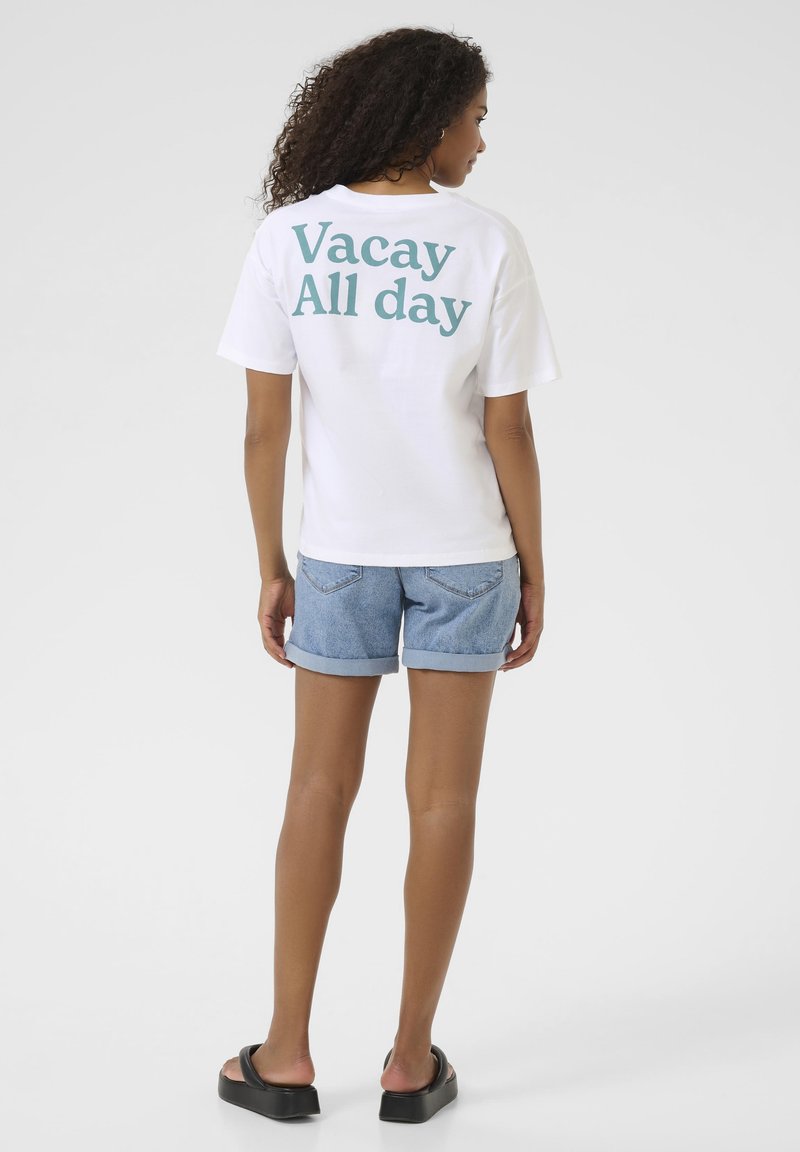 T-shirt en coton blanc avec le texte vert "Vacay All day" au dos, associé à un short en denim bleu. Coupe décontractée et manches courtes.