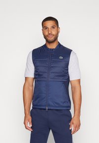 Colete sem mangas azul-marinho com textura acolchoada, fecho éclair completo e bolsos laterais, usado sobre uma camisa de mangas curtas cinza-claro. Detalhe em logo verde.