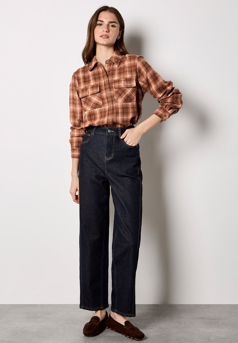 Jeune femme portant une chemise à carreaux marron avec boutons, un jean taille haute foncé, et des mocassins marron, debout contre un mur clair uni.