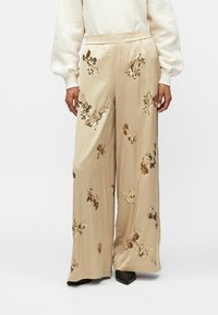 Bež hlače širokega kroja z elastičnim pasom, okrašene s cvetličnim vezenjem z zlatimi in rjavimi okrasnimi sekvincami. Gladka, svilenjasta tekstura tkanine.
