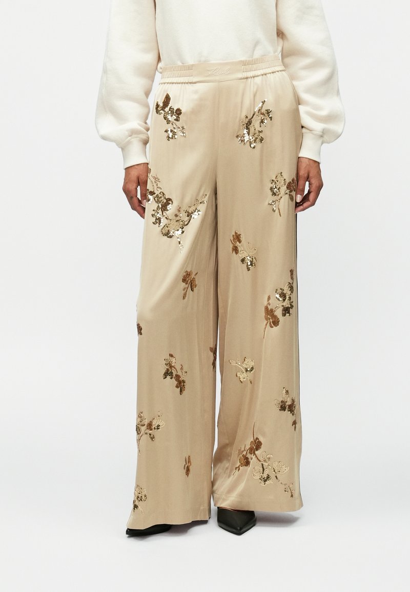 Bež hlače širokega kroja z elastičnim pasom, okrašene s cvetličnim vezenjem z zlatimi in rjavimi okrasnimi sekvincami. Gladka, svilenjasta tekstura tkanine.