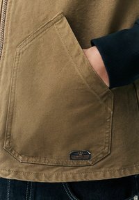 Chaqueta de lona marrón con un gran bolsillo angular, puño de canalé azul marino y una etiqueta en el bolsillo con texto y un logo.