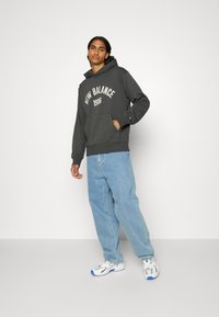 Sudadera gris oscuro con estampado "New Balance 1906", bolsillo delantero tipo canguro, corte relajado, combinada con pantalones vaqueros holgados de color azul claro y zapatillas blancas.