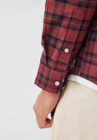 Langärmliges Hemd mit rot-schwarzem Karomuster und Knopfmanschetten; der Stoff scheint strukturiert und leicht zu sein und wird über einem weißen Hemd getragen.