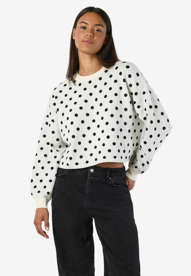 Balts džemperis ar melniem punktiņiem, īsāks dizains, apaļš kakla izgriezums, garas piedurknes, apvienots ar augsto jostu melniem džinsiem.