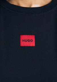 Tamsiai mėlynos spalvos medvilninė marškinėlė, puošti raudonu stačiakampiu etiketė su žodžiu "HUGO" juodai. Klasikinio apvalaus kaklo dizainas.