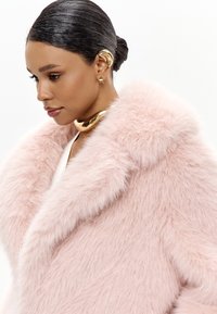 Lunefarvet faux fur frakke med en stor krave og blød tekstur. Modellen bærer et guld choker og unikt guld ørekæde, der fremviser et moderne design.
