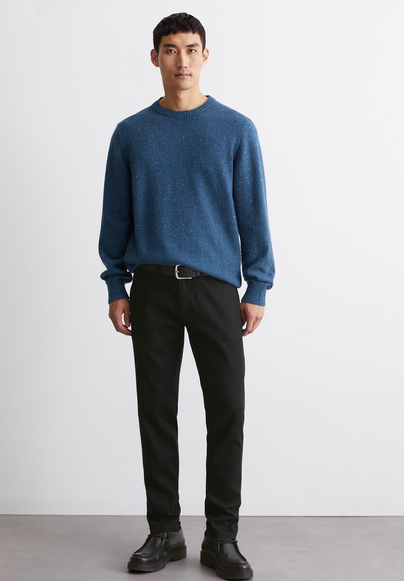 Blau gepunkteter Strickpullover mit gerippten Bündchen, kombiniert mit schwarzen Hosen und schwarzen Schuhen. Das Model steht vor einem neutralen Hintergrund.