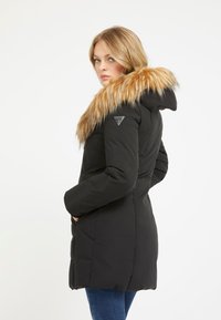 Parka nera isolata con cappuccio in pelliccia sintetica removibile, dotata di una patch con logo sul petto e tasche laterali. Texture liscia e vestibilità su misura.