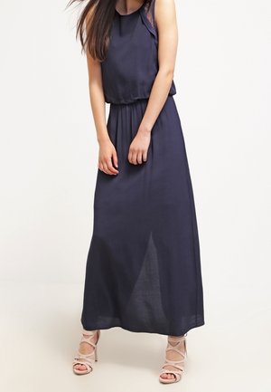 Femme portant une robe maxi bleu marine sans manches avec des détails en dentelle, associée à des sandales à talons haut beige à brides, debout devant un fond uni.