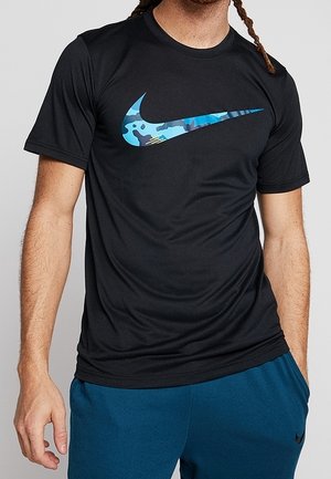 Homme portant un t-shirt noir Nike avec un logo Nike camouflage bleu et un short de sport bleu.