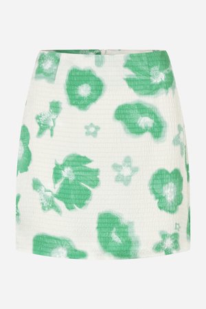 Mini-jupe fleurie blanche avec des motifs de fleurs vertes, en tissu texturé. Elle possède une fermeture éclair dissimulée au dos et une silhouette ajustée.