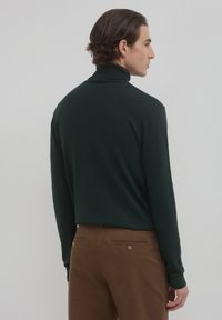 Maglione a collo alto verde scuro realizzato in materiale morbido, con maniche lunghe e una silhouette aderente, abbinato a pantaloni marroni con tasche posteriori.