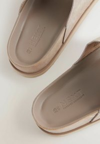 Smeđe mokasine od suedea s glatkom svijetlosmeđom unutrašnjom soles. Sadrži urezanu oznaku "NEXT FOREVER COMFORT" i jednostavan dizajn.