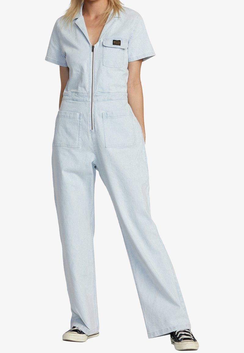 Jumpsuit bleu clair à manches courtes en tissu rayé, avec une fermeture éclair à l'avant, deux poches poitrine et un design à jambes larges. Baskets noires portées.