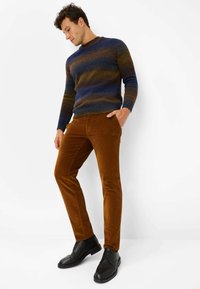 Gestickter Pullover in Blau- und Brauntönen, kombiniert mit schlanken braunen Cordhosen und schwarzen Schnürschuhen. Einfaches, strukturiertes Design.