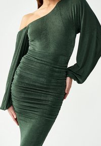 Vihreä, pitkähihainen mekko yhdellä olkapäällä. Sisältää rypytystä vartalon alueella ja sileää, teksturoitua kangasta. Muotoa korostava malli.