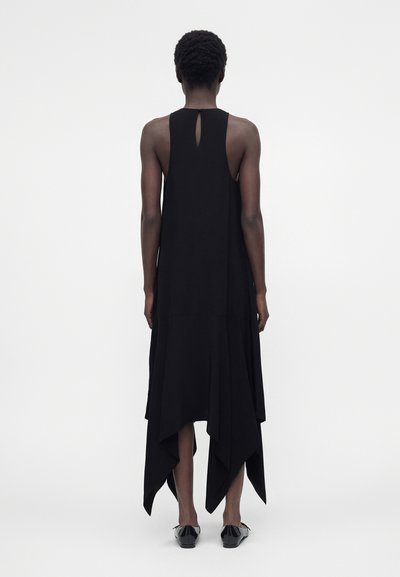 Ψηλό άτομο με κοντά σγουρά μαλλιά φορώντας μαύρο αμάνικο φόρεμα με ασύμμετρη ποδιά και μαύρες μυτερές μπαλαρίνες, στέκεται με την πλάτη προς το μέρος μας.