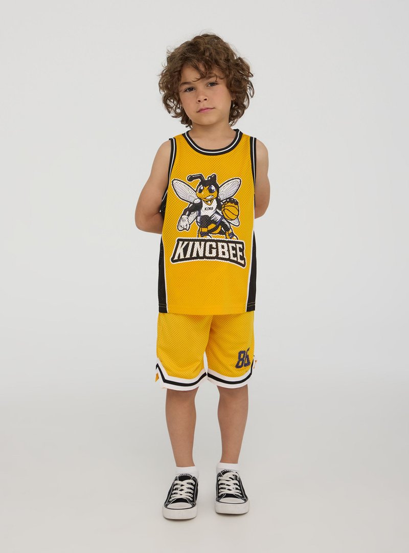 Bambino con capelli ricci che indossa un completo da basket giallo con un logo di ape e scarpe da ginnastica nere, in piedi davanti a uno sfondo semplice.