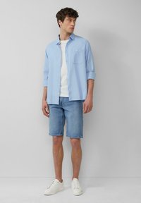 Lichtblauw knoopshirt met opgerolde mouwen, een wit T-shirt eronder, denim shorts en witte sneakers. Eenvoudige, casual outfit.