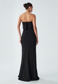 Nainen, jolla on hiukset nutturalla, pukeutunut olkaimettomaan mustaan iltapukuun ja kullanvärisiin korvissilmiin ja rannekoruun, seisoo tasaisen vaaleataustan edessä.