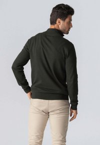 Mørkegrøn pullover med krave og lange ærmer, lavet af et glat strikket stof. Båret over lyse beige bukser, viser bagsiden.