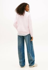 Donna con capelli ricci che indossa un maglione rosa chiaro, una camicia a righe, jeans larghi blu e sneaker bianche, in piedi contro uno sfondo bianco.