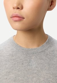 Ljusgrå tröja med ribbad halsringning och ett broderat blomlogotypdetalj på bröstet. Mjuk textur med en klassisk passform.