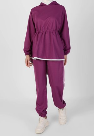 Femme portant un survêtement à capuche violet avec taille et poignets élastiques, bordure blanche et baskets blanches sur un fond gris clair.