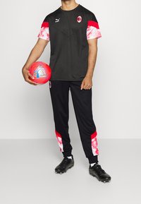 Puma träningskläder i svart med röda och rosa tie-dye-accenter; lätt T-shirt och avsmalnande byxor; svarta fotbollsskor; håller en klar röd fotboll.
