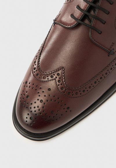 Ruskea nahkainen kenkä, jossa on rei'itetyt yksityiskohdat, siipikuviointi, pyöreä kärki ja kontrastivärinen musta pohja teksturoidulla pinnalla.