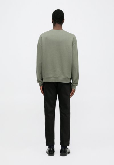 Groene oversized sweatshirt met geribbelde manchetten, gecombineerd met zwarte broeken en zwarte schoenen. De textuur is glad en de pasvorm is ontspannen.