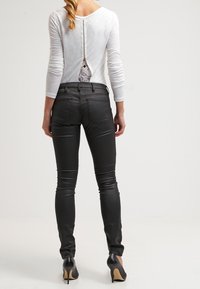 Top blanco de manga larga con detalle de encaje en la espalda y pantalones ajustados negros. La modelo lleva tacones altos negros. El conjunto cuenta con un cierre de cremallera.