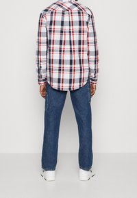 Gekleurde shirt met een ruitpatroon in marineblauw, rood en wit, met knopenmanchetten. Gemaakt in combinatie met blauwe spijkerbroeken en witte sneakers. Zicht vanuit de achterkant.