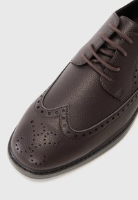 Zapato de vestir de cuero marrón con detalles perforados, puntera redonda y cordones. La textura es suave con un acabado ligeramente brillante. Diseño casual pero refinado.