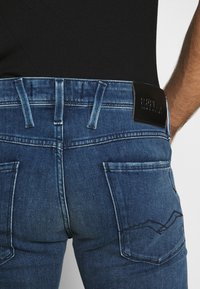 Vaqueros de denim en azul oscuro con una textura descolorida. Presentan dos bolsillos traseros y una etiqueta de cuero negra de marca en la cintura.