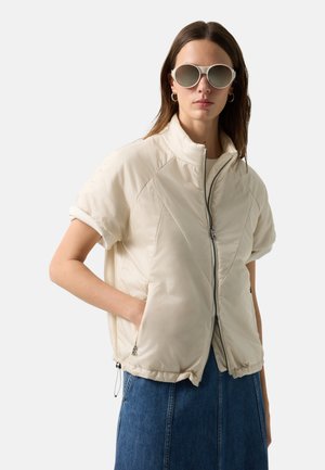 Kobieta ubrana w oversize'ową beżową puchową kurtkę z krótkim rękawem, niebieską spódnicę dżinsową, duże okrągłe okulary przeciwsłoneczne i kolczyki-koła na białym tle.