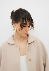 Cappotto di lana beige con colletto ampio e chiusura a un bottone. Orecchini a cerchio spessi e curvi in oro. La modella ha i capelli scuri e mossi.
