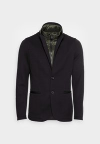 Svart strukturerad kavaj med en grön pufferkrage. Har två framfickor och en enknappsstängning. Materialet verkar vara slätt och texturerat.